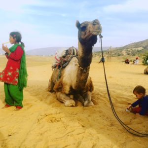 Kind spielt im Sand, daneben entspannt ein Kamel